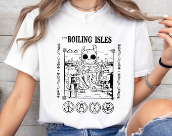 May include: White t-shirt featuring a black and white graphic design from "The Boiling Isles". The design includes a skull-like creature, buildings, and symbols. The text "THE BOILING ISLES" is at the top of the design.