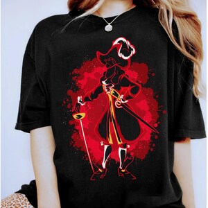May include: Black t-shirt featuring a red, yellow and white graphic of Captain Hook from Peter Pan. The graphic is in a watercolour style with splashes of red paint.