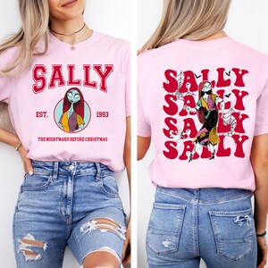 K&ouml;nnte beinhalten: Ein pinkfarbenes T-Shirt mit einem roten "Sally"-Motiv auf der Vorderseite. Das Motiv enth&auml;lt ein Cartoon-Bild von Sally aus dem Film "Nightmare Before Christmas" und den Text "EST. 1993" und "THE NIGHTMARE BEFORE CHRISTMAS". Die R&uuml;ckseite des Shirts hat ein rotes "Sally"-Motiv, das f&uuml;nfmal wiederholt wird, mit einem Cartoon-Bild von Sally in der Mitte.