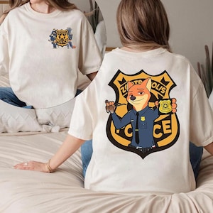 Puede incluir: Camiseta de color crema con una insignia de polic&iacute;a de dibujos animados en la parte delantera y un dise&ntilde;o m&aacute;s grande en la parte trasera. El dise&ntilde;o trasero incluye un personaje de zorro con uniforme de polic&iacute;a, sosteniendo una placa, con el texto "Zootopia Police".