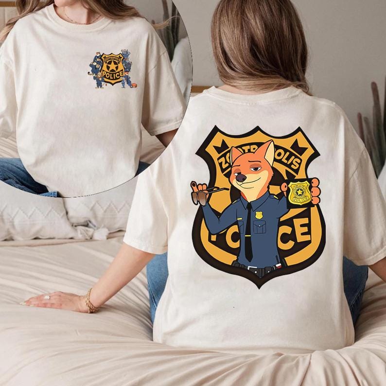 Puede incluir: Camiseta de color crema con una insignia de polic&iacute;a de dibujos animados en la parte delantera y un dise&ntilde;o m&aacute;s grande en la parte trasera. El dise&ntilde;o trasero incluye un personaje de zorro con uniforme de polic&iacute;a, sosteniendo una placa, con el texto "Zootopia Police".