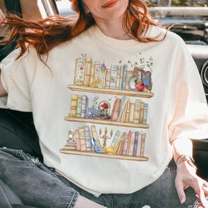 Può includere: Una maglietta bianca con una grafica di una libreria piena di libri, una rosa, una scarpetta di vetro, un candelabro e una mela. I libri sono di diversi colori e dimensioni.