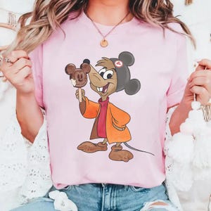 Puede incluir: Camiseta rosa claro con un personaje de dibujos animados sosteniendo un helado con forma de Mickey Mouse. El personaje lleva una chaqueta naranja, una camisa roja y un sombrero gris con orejas negras. El helado es marr&oacute;n.