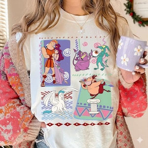Puede incluir: Camiseta blanca con gráficos que presenta ilustraciones coloridas de personajes de una película animada popular. El diseño incluye a Hércules y Megara, Hades, Pegaso y otros personajes. La camiseta tiene cuello redondo y mangas cortas.