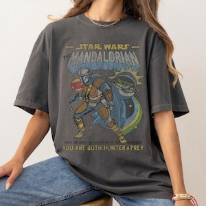 K&ouml;nnte beinhalten: Ein dunkelgraues T-Shirt mit einem Grafikdruck eines Charakters aus Star Wars: The Mandalorian. Der Text auf dem Shirt lautet "Star Wars - The Mandalorian" und "Where you choose to be is the way of the Mandalorian. You are both hunter & prey."