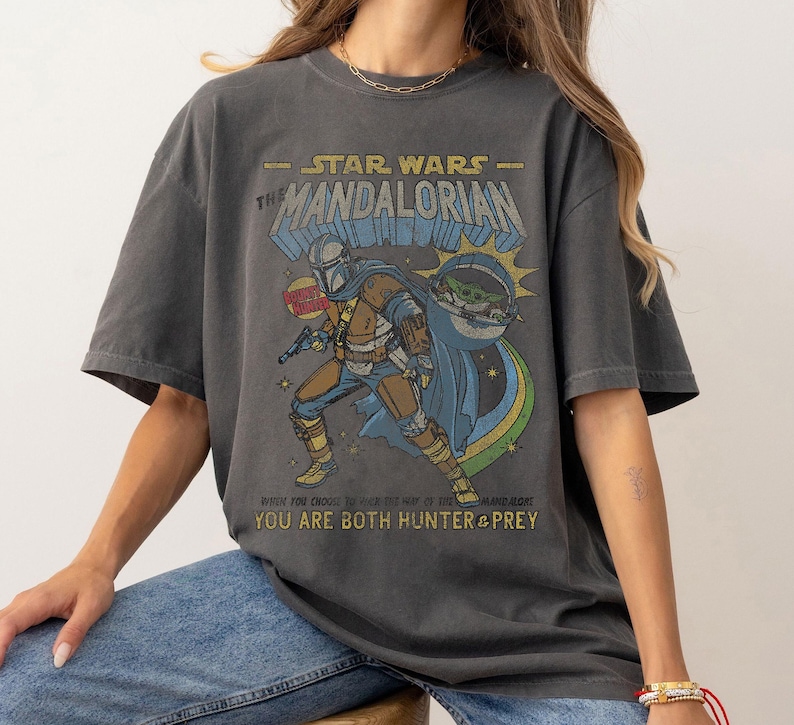 K&ouml;nnte beinhalten: Ein dunkelgraues T-Shirt mit einem Grafikdruck eines Charakters aus Star Wars: The Mandalorian. Der Text auf dem Shirt lautet "Star Wars - The Mandalorian" und "Where you choose to be is the way of the Mandalorian. You are both hunter & prey."