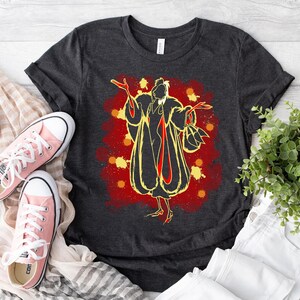 May include: A dark grey t-shirt featuring a graphic print of Cruella De Vil in a yellow fur coat with red accents. The background is a red and orange splatter pattern.
