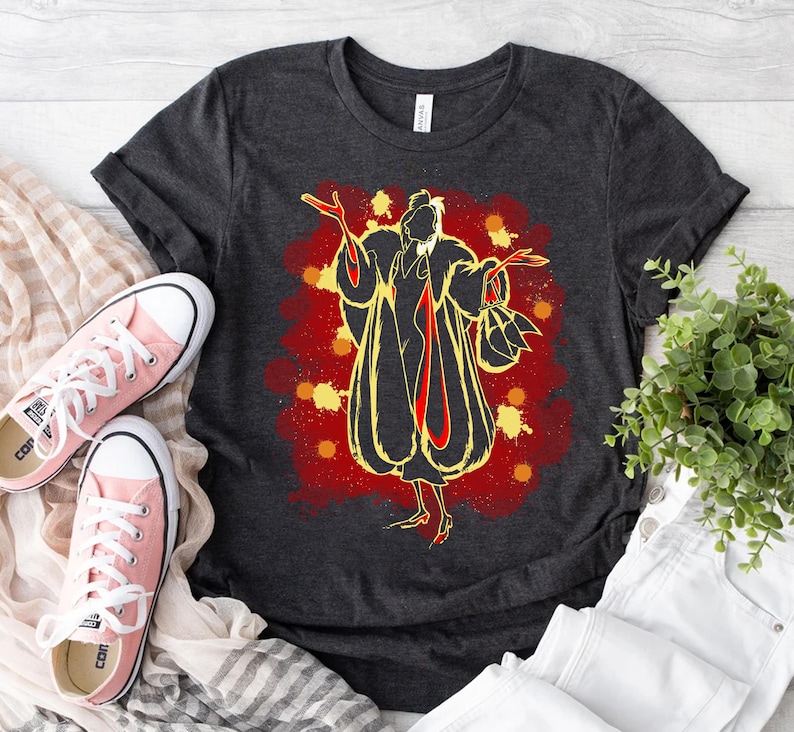 May include: A dark grey t-shirt featuring a graphic print of Cruella De Vil in a yellow fur coat with red accents. The background is a red and orange splatter pattern.