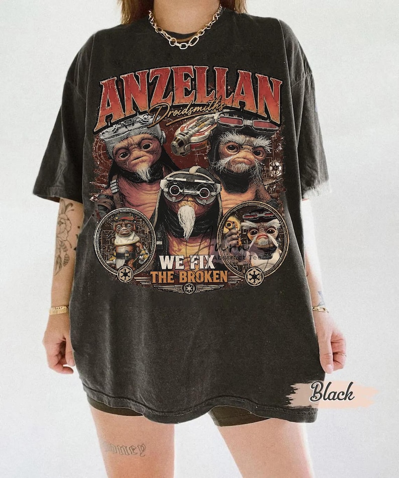 May include: A black short-sleeve t-shirt featuring a graphic print of three Anzellan characters with goggles. The text reads "ANZELLAN Droidsmiths" and "WE FIX THE BROKEN" with small circular emblems. The shirt has a faded, washed-out appearance.