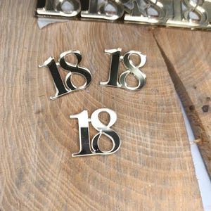 Può includere: Decorazioni dorate con il numero 18 e contorno nero, sparse su una superficie di legno. Altre sono visibili sullo sfondo. Questi numeri decorativi sono adatti per feste di compleanno o altri eventi speciali.