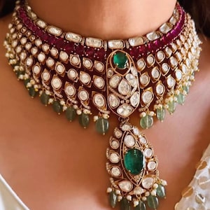 Puede incluir: Un collar de oro ornamentado con un gran colgante de esmeralda y pequeños detalles de esmeraldas y perlas. El collar tiene un diseño en capas con piedras tipo rubí y diamante. Una joya lujosa.
