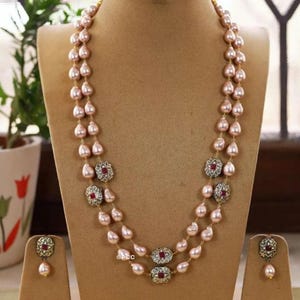 Op de afbeelding: Een ketting met twee strengen en bijpassende oorbellen. De ketting heeft roze druppelvormige parels met sierlijke goudkleurige accenten en rode edelstenen. De oorbellen hebben een enkele roze parel en een bijpassend goud- en rood edelsteenontwerp.