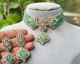 Op de afbeelding: Een driedelige sieradenset met een choker-ketting, hanger en oorbellen. De ketting en oorbellen hebben smaragdgroene stenen en zijn versierd met kleine, heldere kristallen. De hanger heeft een druppelvorm. De sieraden worden getoond op een grijze mannequinhoofd.