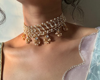 May include: A gold-toned choker necklace with multiple strands of small, white pearls. The necklace features dangling gold pendants with clear and pearl-like accents. The jewelry is worn on a person.