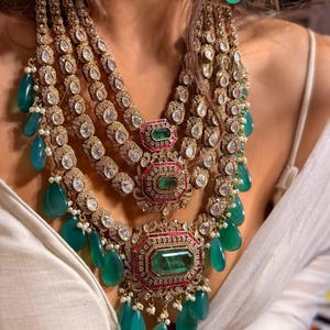May include: An elaborate gold-toned necklace and matching earrings set. The necklace features multiple layers of clear, sparkling stones and large emerald-green teardrop-shaped gems. The earrings have a similar design. The jewelry is set against a white background.