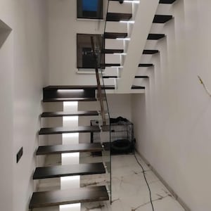 Może przedstawiać: Nowoczesne schody z ciemnymi drewnianymi stopniami i białymi podstopniami. Schody mają szklaną balustradę i są oświetlone diodami LED. Schody znajdują się w białym pokoju z białą podłogą.