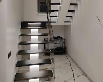 May include: A modern staircase with dark wood steps and white risers. The staircase has a glass railing and is lit by LED lights. The staircase is in a white room with a white tiled floor.