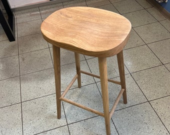 May include: A light brown wooden stool with a smooth, oval-shaped seat and four tapered legs connected by a square support frame. The wood grain is visible across the surface of the seat and the legs, showcasing a natural light tan finish.