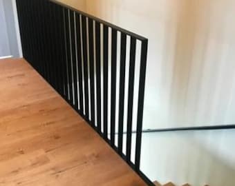 May include: Black metal railing with vertical bars, installed on a wooden staircase. The railing is attached to a wall with a light brown wooden floor.
