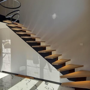 Może przedstawiać: Nowoczesne schody z drewnianymi stopniami i czarnymi metalowymi balustradami. Schody są oświetlone światłem wpuszczonym w sufit i znajdują się w pomieszczeniu z białymi ścianami i marmurową podłogą.