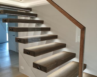 May include: A floating staircase featuring thick, dark brown wooden treads with integrated LED lighting underneath each step. A clear glass panel serves as a side railing, supported by a thin, angled dark brown wooden handrail that follows the stairs.