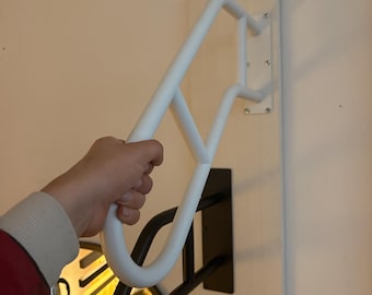 May include: A white metal wall-mounted coat rack with a unique, curved design. The rack is attached to a light-colored wooden board, with a black metal support underneath. The rack is being held by a person's hand.