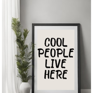 以下が含まれることがあります： 白い背景に黒い文字で「COOL PEOPLE LIVE HERE」と書かれた、黒い額縁の壁掛けアートです。