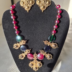 May include: A gold-coloured necklace and matching earrings set. The necklace features a strand of red beads, gold filigree accents, and colourful glass beads. The earrings have a similar gold filigree design. The jewellery is displayed on a black velvet bust.