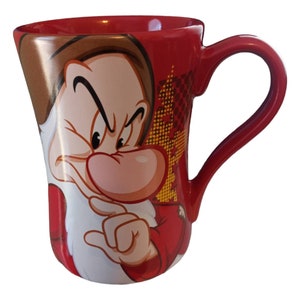 Puede incluir: Taza de cerámica roja con una ilustración de Gruñón de Blancanieves. La taza tiene forma curva y un asa roja. El diseño incluye la cara de Gruñón y un fondo rojo y amarillo.