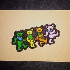 May include: An embroidered patch featuring four dancing bears in a row. The bears are colored green with a blue collar, yellow with a green collar, orange with a green collar, and white with a blue star, all outlined in black thread on a dark background.