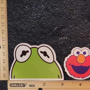 May include: Two stickers featuring cartoon characters. One is a green frog with large white-rimmed eyes. The other is a red character with an orange nose and purple mouth. The stickers are approximately 2 inches wide.
