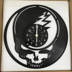 May include: A black vinyl record clock featuring a Grateful Dead Steal Your Face skull design. The clock face is a smaller circle with white numbers and hands.