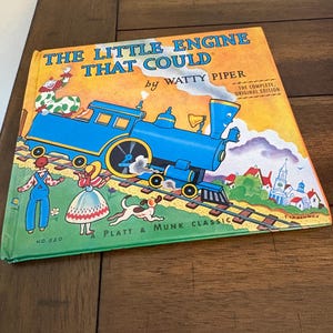 May include: A vintage children's book titled "The Little Engine That Could" by Watty Piper. The cover features a blue train, illustrations of children, a dog, and a town. The book is a Platt & Munk Classic.