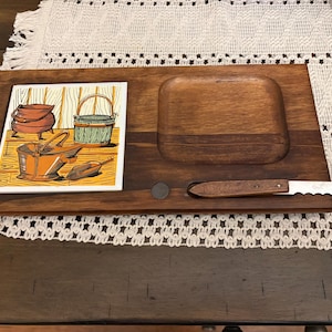 May include: Wooden serving board with a recessed area, a decorative tile, and a serrated knife. The tile features illustrations of vintage buckets and a pot. The board is made of dark wood.