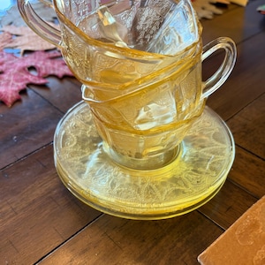 May include: A stack of three yellow glass teacups with matching saucers. The cups and saucers have a detailed embossed pattern. The cups have handles and are stacked on top of each other. The set is on a wooden table.