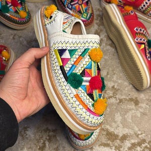 Op de afbeelding: Witte leren instappers met kleurrijke geborduurde geometrische patronen en pompom accenten. De schoenen hebben een dikke, gelaagde zool met een lichtbruine rand. Andere paren schoenen met vergelijkbare ontwerpen zijn zichtbaar.