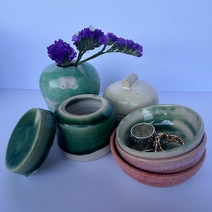 Puede incluir: Una colección de cerámica en tonos verdes, beige y rosa. El arreglo incluye un pequeño jarrón con flores moradas, un tarro con tapa, un pequeño tarro abierto y una pila de cuencos. Un anillo de plata y joyas de oro están en uno de los cuencos.
