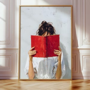 May include: A framed art print showing a person with a red book covering their face. The person wears a white shirt. The artwork is set against a light grey background and framed in a gold-coloured frame. The image is in a room with wooden flooring.