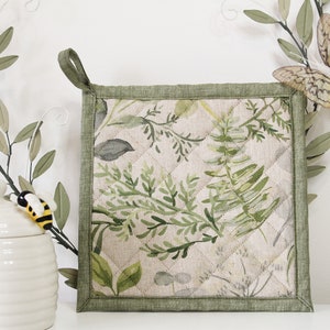 May include: A green quilted pot holder with a floral pattern featuring leaves and ferns. The pot holder has a green border and a loop handle.