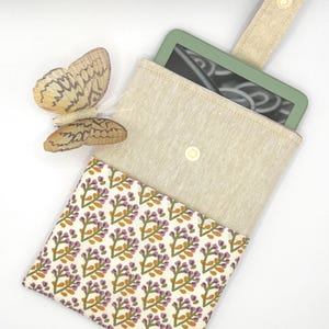 Puede incluir: Una funda beige de tela para lector de libros electrónicos con solapa, con un estampado floral en morado, verde y dorado. La funda contiene un lector de libros electrónicos verde. Una decoración de mariposa beige está en el lado izquierdo.