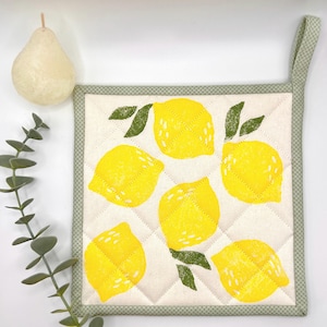 Puede incluir: Un salvamanteles cuadrado y acolchado con un patrón repetido de limones amarillos y hojas verdes sobre un fondo claro. Tiene un borde verde y un lazo para colgar. Mide aproximadamente 20 cm x 20 cm.