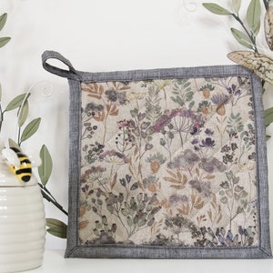 May include: A grey and white floral print pot holder with a grey loop handle. The pot holder is made of fabric and has a quilted design.