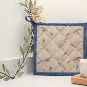 May include: A blue and beige quilted pot holder with a seagull pattern and a denim trim.