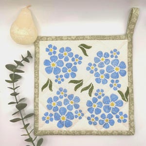 May include: A square quilted pot holder featuring a pattern of blue flowers and green leaves on a cream background. It has a green floral border and a hanging loop. A pear-shaped candle and eucalyptus sprig are in the background.