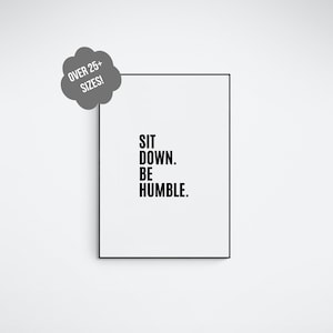 May include: A black and white framed print with the text "SIT DOWN. BE HUMBLE." on a white wall. The print is hanging above a white floor with a silver metal rope barrier.