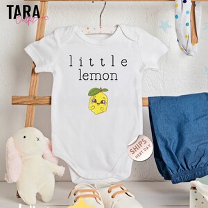 May include: White baby bodysuit with the text "little lemon" and a cartoon lemon graphic. The outfit includes blue denim pants, white shoes, and a plush bunny. A headband and a wooden toy are also visible.