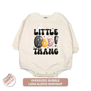May include: Cream-colored, long-sleeved baby bodysuit with the text "LITTLE BOO THANG" in black, with a ghost and colorful donut-shaped letters. The bodysuit has snap closures and a round neckline. A banner at the bottom reads "OVERSIZED BUBBLE LONG-SLEEVE BODYSUIT".