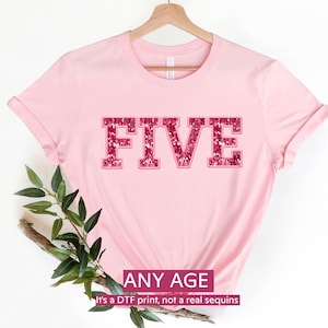 May include: A light pink t-shirt with the word "FIVE" in a sequined font. The text "ANY AGE" is below the word "FIVE". The shirt is hanging on a wooden hanger.