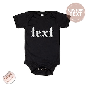 May include: Black baby bodysuit with the word "text" in white gothic font. The bodysuit has short sleeves and snaps at the bottom. A circular sticker with the words "Custom Text" is in the upper right corner.