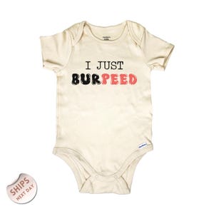 May include: Cream-colored baby bodysuit with short sleeves. The text "I JUST BURPEED" is printed across the front in black and coral letters. The bodysuit has a snap closure at the bottom and a round neckline.